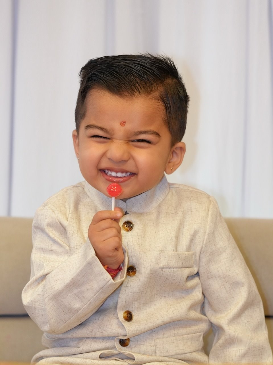 A young boy in a cream suit grinning with a lollipop