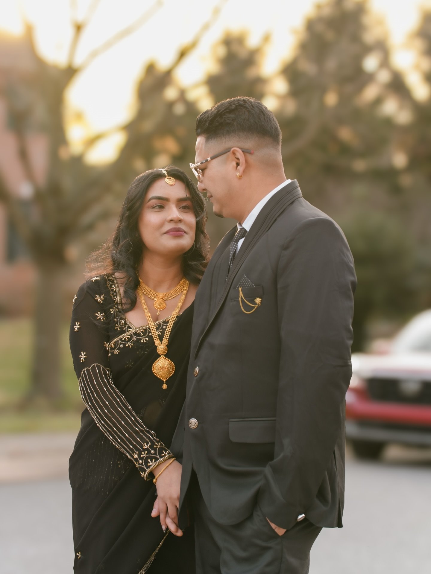 A couple in black formal wear standing close at golden hour, she looks up at him