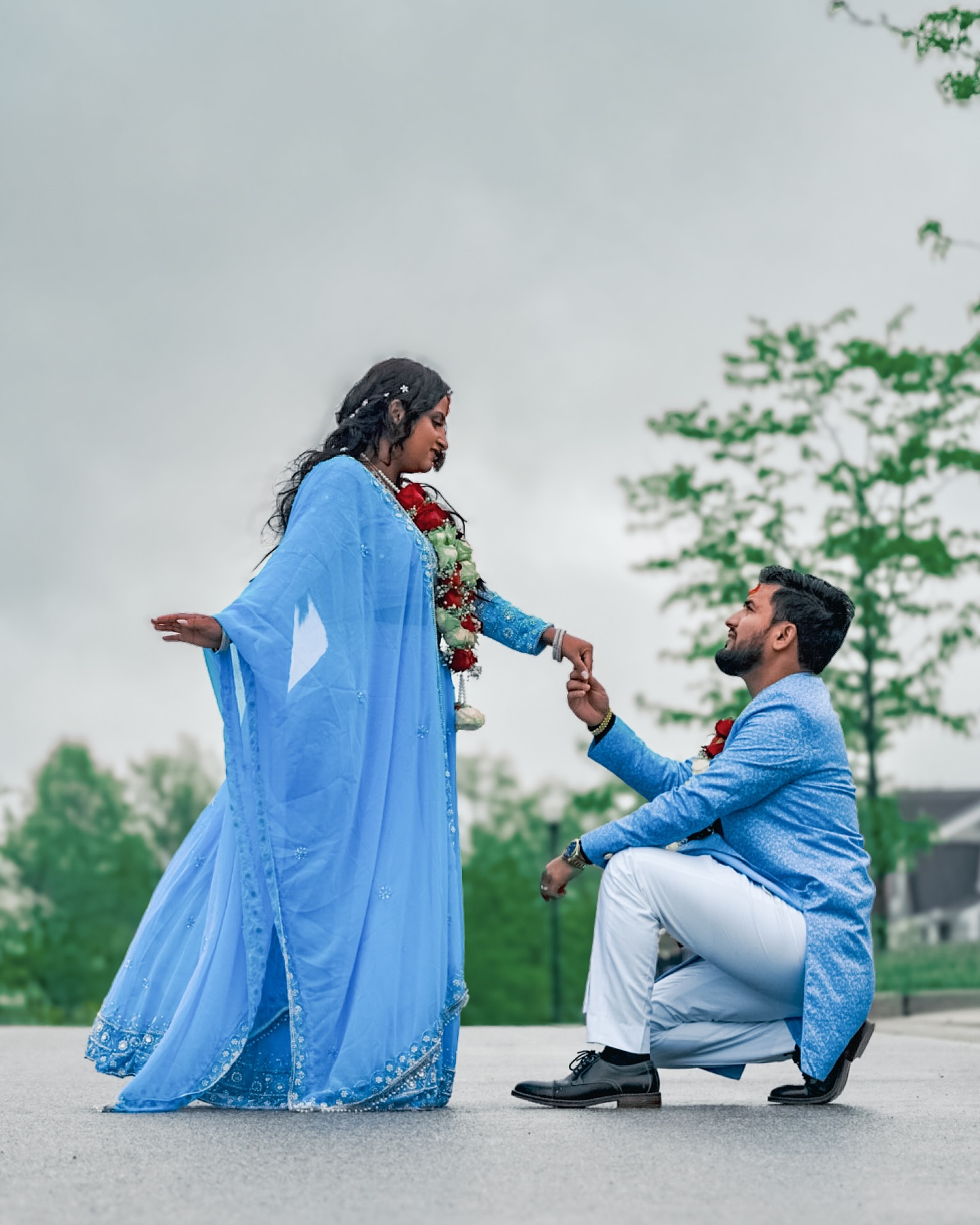 A man in a blue kurta on one knee, holding the hand of a woman in a blue saree