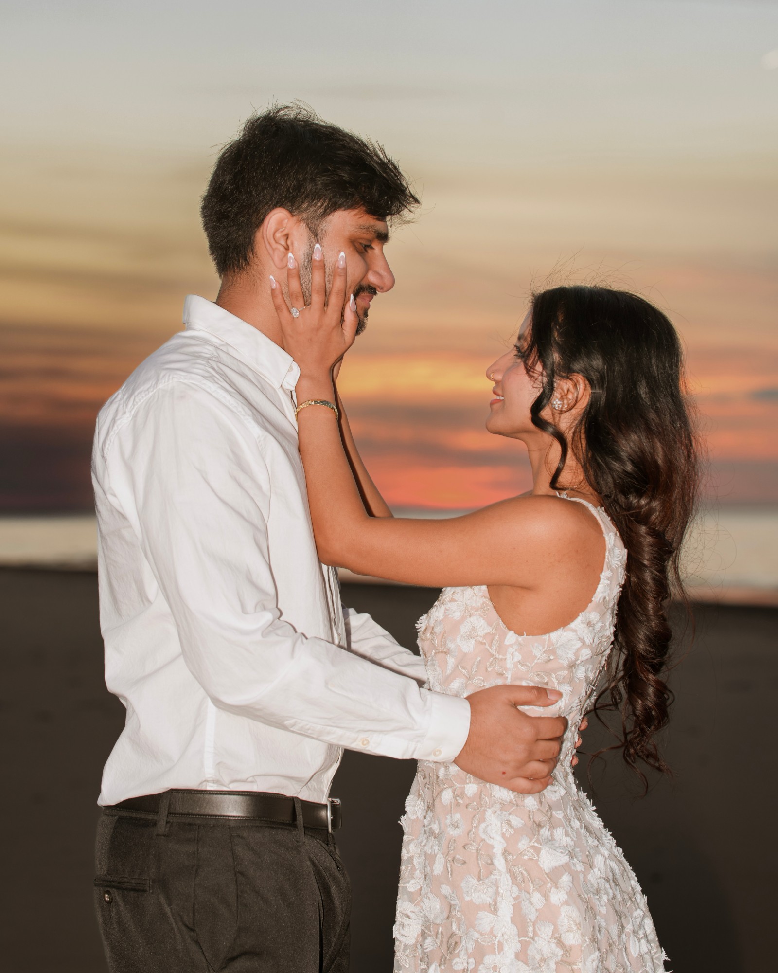 A couple on the beach at sunset, her hand on his cheek as they smile at each other