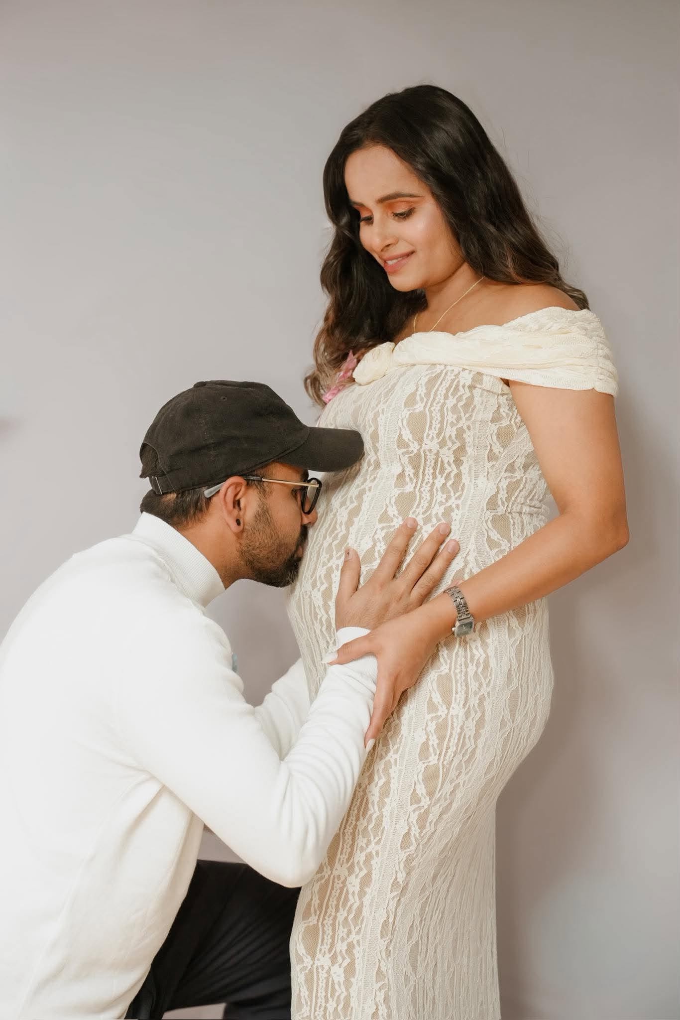 An expectant mother in a cream lace dress smiling down as her partner kneels to kiss her bump