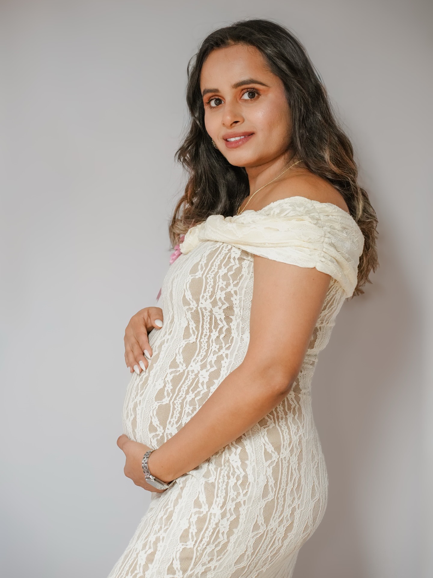 An expectant mother in a cream lace dress holding her bump and smiling at the camera
