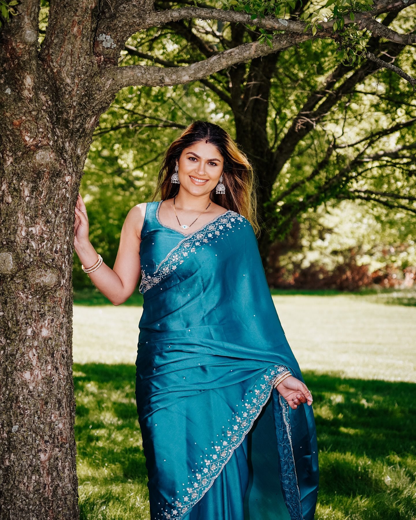 A woman in a teal saree standing beside a tree in a sunlit park