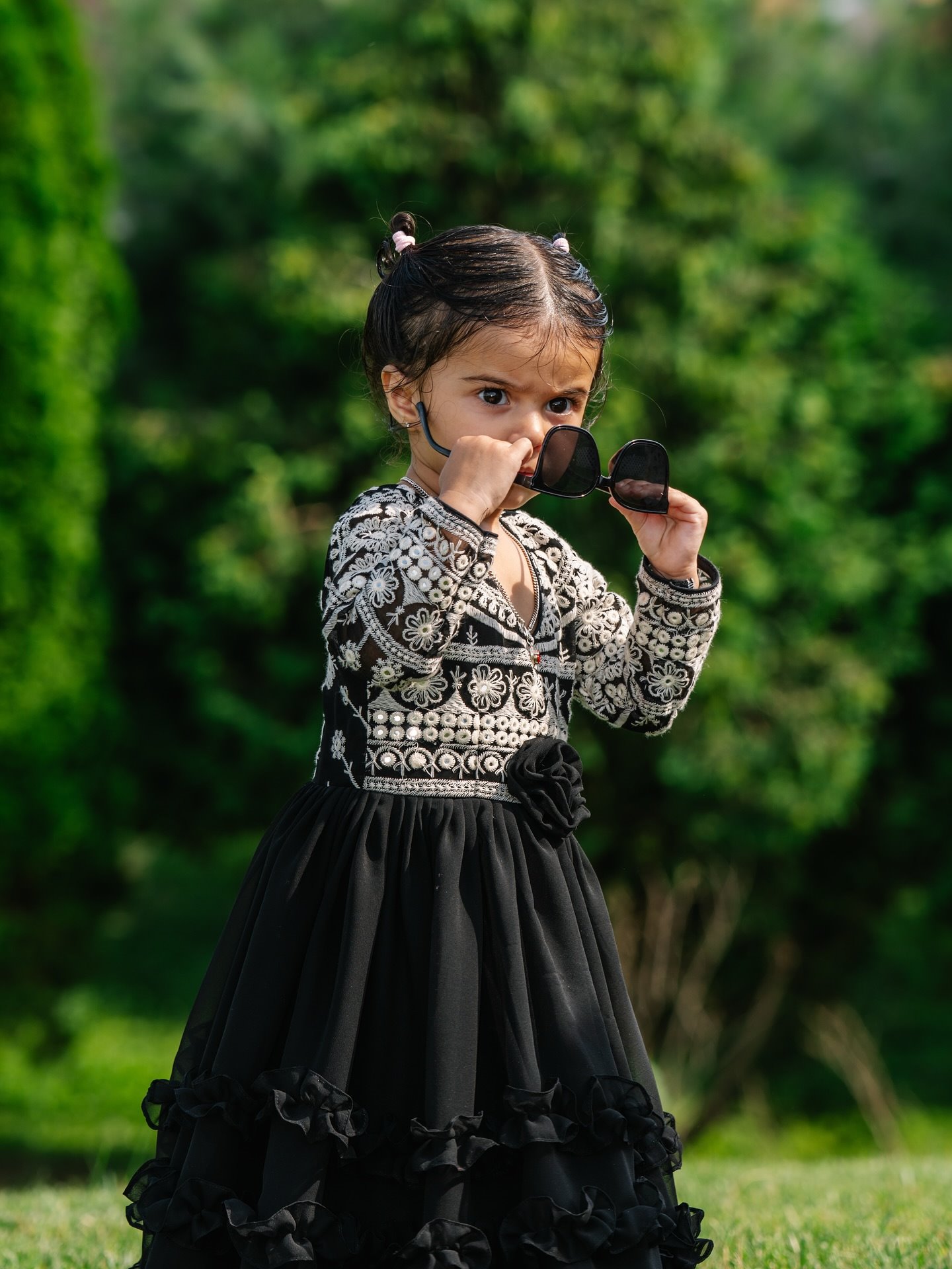 A toddler in an embroidered black dress pulling down a pair of sunglasses