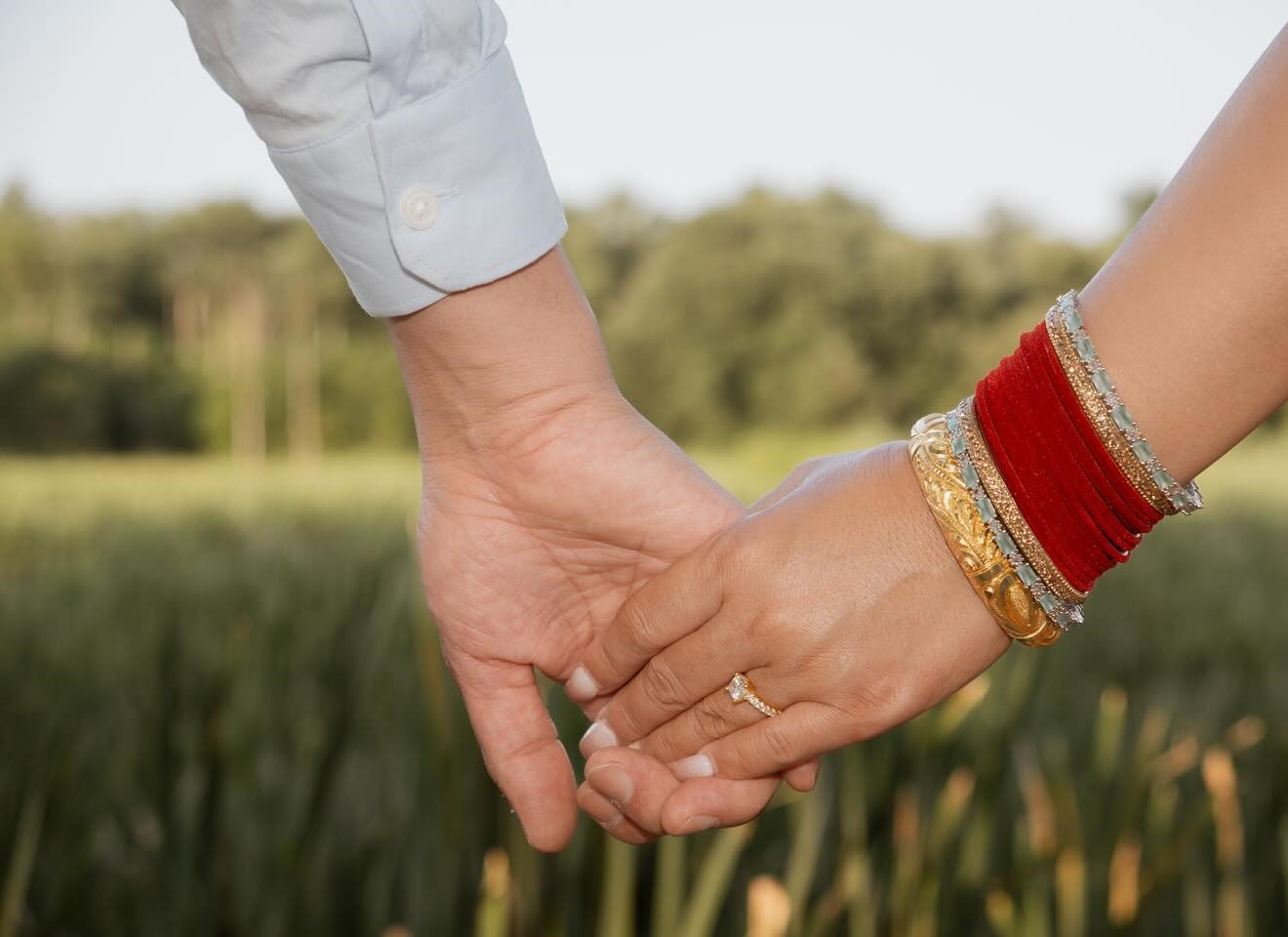 A bride and groom holding hands in front of a field, her wrist stacked with red and gold bangles